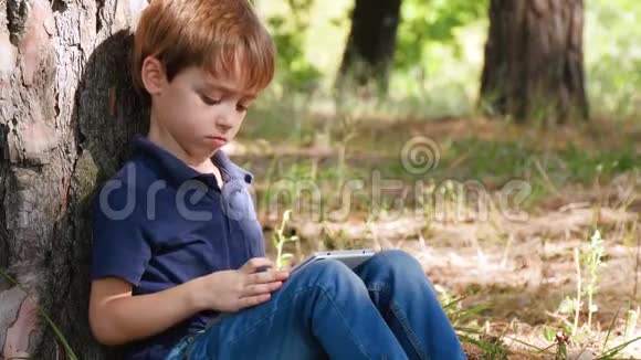 学龄前小男孩的肖像一个孩子坐在一棵树旁看着平板电脑上的东西视频的预览图