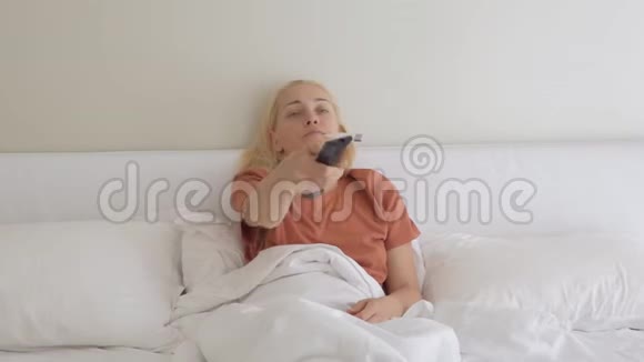 这位女士躺在家里的床上用电子设备切换遥控电视频道测量她的体温视频的预览图