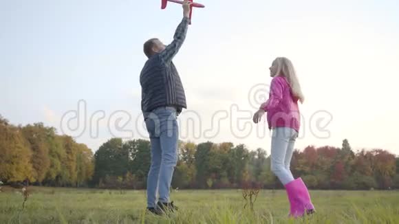 年轻的白种人父亲在秋天的草地上和他十几岁的金发女儿玩耍持有粉红色玩具飞机的成年男子视频的预览图