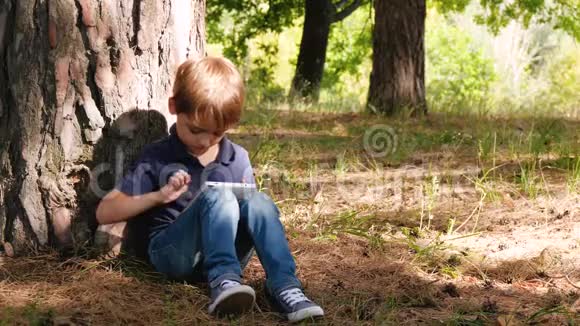 可爱的男孩坐在公园的一棵树旁快乐的孩子热情地看着平板电脑的屏幕触摸着屏幕视频的预览图