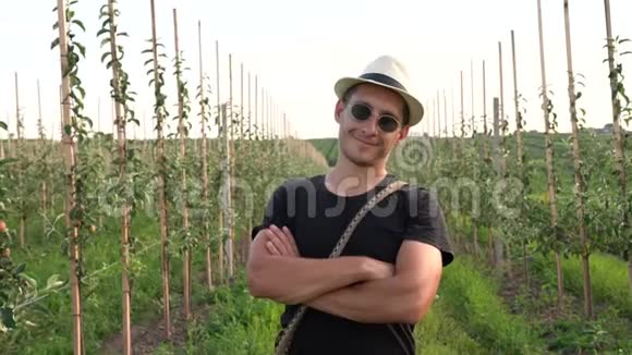 穿着黑色T恤太阳镜和帽子的快乐青年站在他的苹果花园里双手戴着胸衣看着视频的预览图