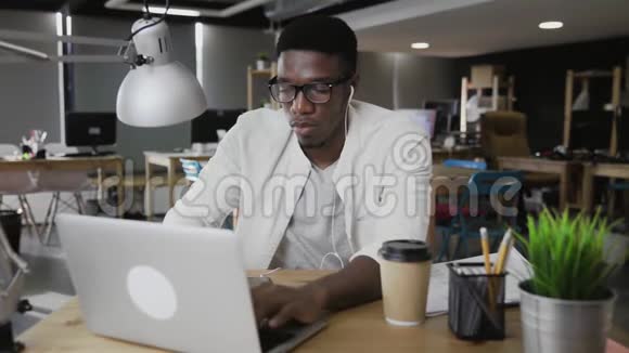 带耳机的非洲商人用笔记本电脑在现代办公室看文件视频的预览图