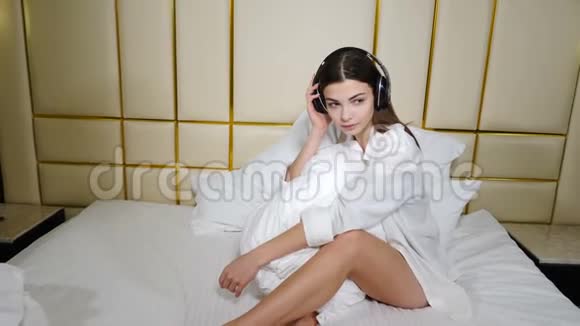 美丽的年轻女子听着音乐微笑着拿着耳机躺在家里的床上技术青年视频的预览图