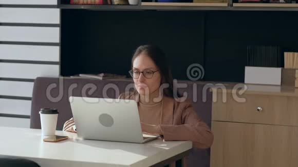 戴眼镜的漂亮女人在咖啡店里使用笔记本电脑和喝咖啡视频的预览图