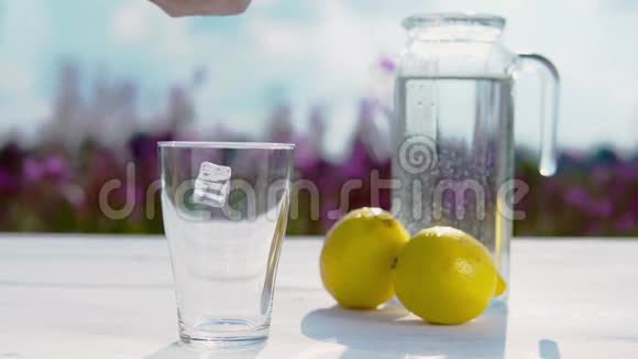 手把冰块扔在一个空杯子里站在白色的桌子上对着一个带水和两个柠檬的装饰器视频的预览图