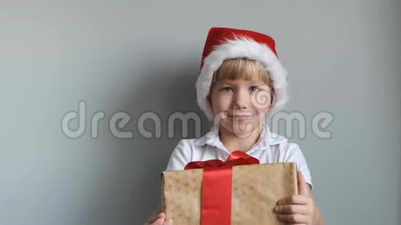 戴着圣诞帽的可爱男孩戴着灰色背景的礼盒看着相机微笑着视频的预览图