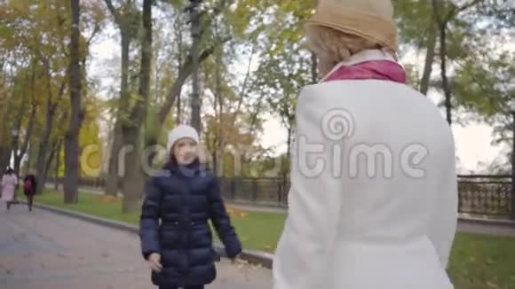 可爱的白种人女孩戴着白色的帽子和温暖的蓝色外套和她的祖母在公园度过了秋天的一天成熟的女人视频的预览图