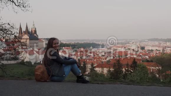 美丽的快乐女孩带着背包坐在山上的轨道上有着古老的布拉格的风景如画的景色视频的预览图