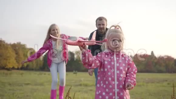 小可爱的金发女孩在秋天森林的背景下带着粉红色的玩具飞机奔跑她的父亲和姐姐视频的预览图