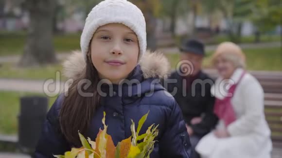 微笑着戴着白帽子的白种人女孩手里拿着一束黄叶看着镜头她的祖父母视频的预览图