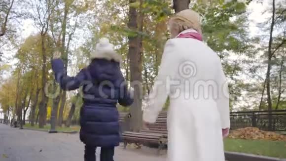 一个戴着白帽子穿着暖蓝色外套的白种人女孩在她祖母面前蹦蹦跳跳地沿着小巷走着视频的预览图