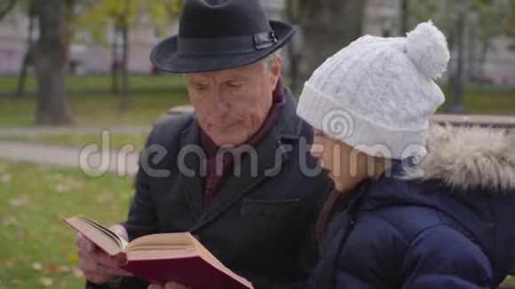 一个穿着经典衣服的白种人成熟男子和孙女坐在长凳上读一本书视频的预览图