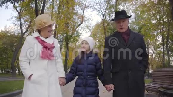穿着白色帽子和蓝色外套的漂亮白人女孩和她的祖父母在公园的小巷里散步高级视频的预览图