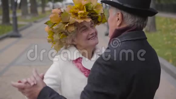 戴着黄叶花环的白种人成熟的女人微笑着和她可爱的丈夫在小巷里跳舞视频的预览图