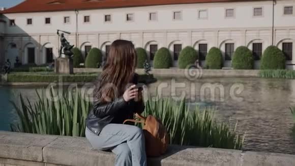 年轻迷人的女孩在池塘边美丽的公园里坐着智能手机有瓦伦斯坦宫和视频的预览图