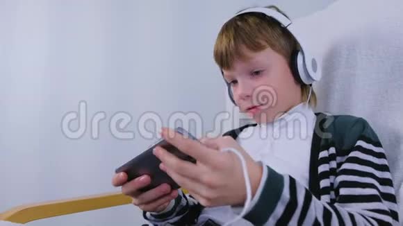 快乐男孩正坐在家里的扶手椅上玩智能手机耳机游戏视频的预览图