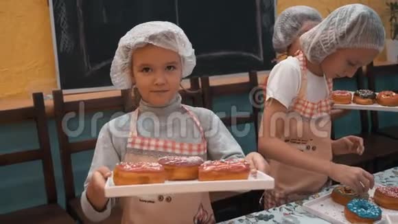 快乐女孩戴着厨师帽和围裙在烹饪大师班上展示带有釉面甜甜圈的托盘带烤的可爱厨师女孩视频的预览图