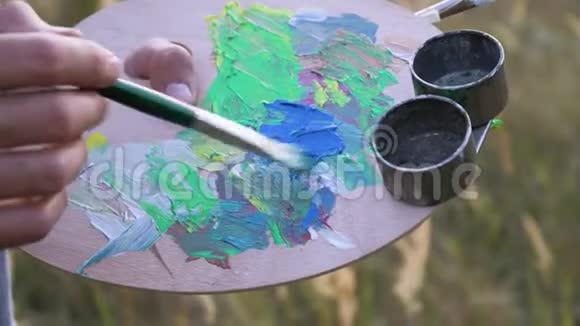 男性艺术家在自然景观中的空地上将调色板上的颜色混合在一起调色板和画笔的特写视图视频的预览图