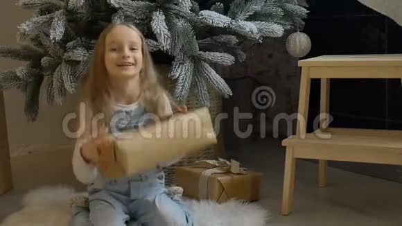 可爱的金发女孩收到了一个礼品盒里面包着工艺纸坐在圣诞树附近快乐的童年和圣诞节视频的预览图