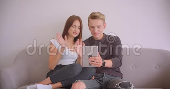 一对年轻可爱的白种人夫妇在平板电脑上打了个视频电话他们坐在平板电脑上挥手视频的预览图