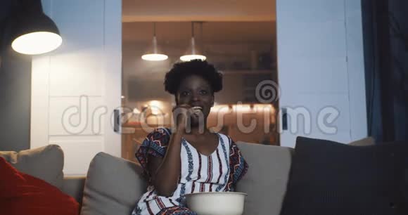 快乐的年轻千禧非洲女人微笑着在家里的沙发上看电视肥皂剧带着零食慢动作视频的预览图