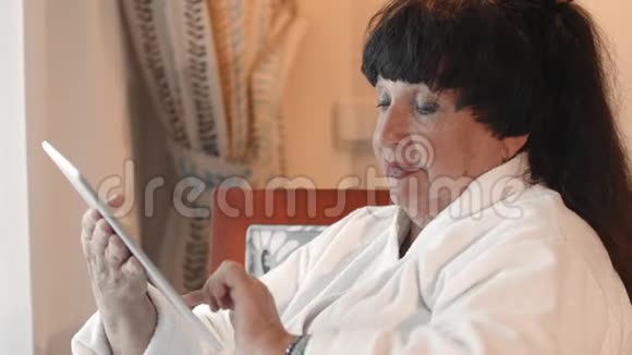 一位穿着白色特里长袍的快乐微笑的白人高级妇女正在使用平板电脑写着一条信息使用的概念视频的预览图