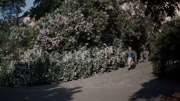 穿着皮夹克牛仔裤和靴子的可爱女孩在春天的公园里在一堵开花的灌木丛的墙上散步视频的预览图