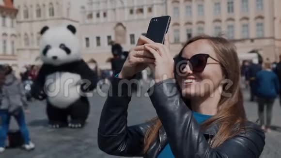 戴太阳镜的漂亮女孩站在城镇广场上拍摄历史建筑一个穿着熊猫服装的男人视频的预览图