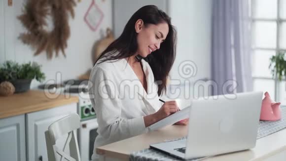 穿着浴袍的年轻女子坐在厨房在笔记本上做笔记侧视视频的预览图