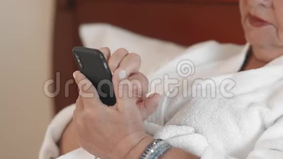 穿着白色浴袍使用智能手机输入聊天信息的未被认出的成年女性使用技术和视频的预览图