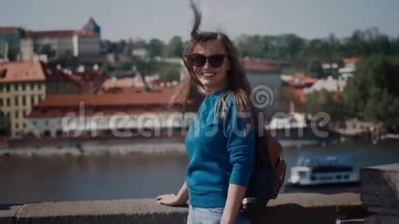 微笑的年轻女子戴着太阳镜站在河边的露台上她的头发在大风中飘扬视频的预览图