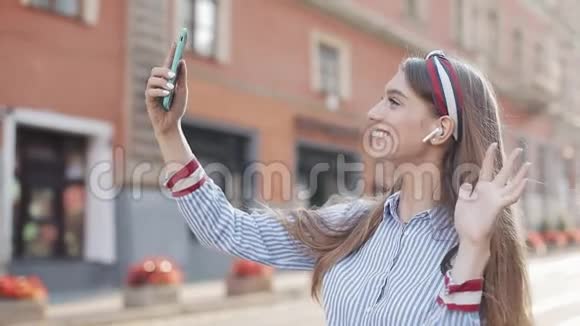 穿着带条纹衬衫和耳机的棕色头发和时髦头巾的漂亮年轻女孩视频通话视频的预览图