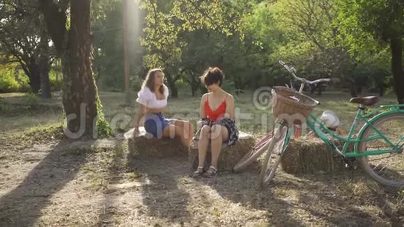 两个可爱的女人在夏天的花园里坐在干草堆上聊天她们的自行车站在附近农村生活复古视频的预览图