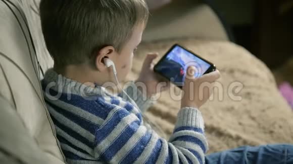 一张可爱的金发小男孩拿着一部智能手机在灰色沙发上打着枕头的肖像视频的预览图