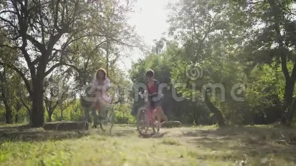 两个快乐的女朋友在夏天的公园或花园里骑自行车从摄像机旁边经过的女孩农村生活复古风格视频的预览图