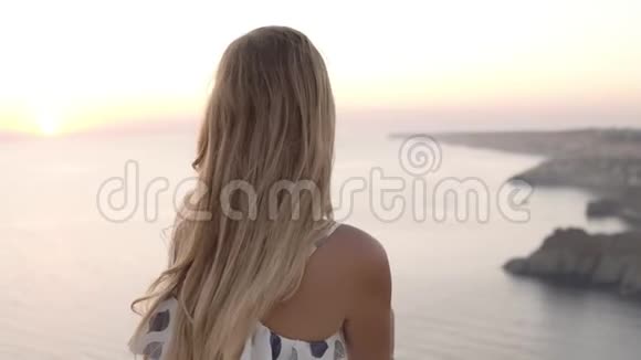 一个头发长长的金发女郎正坐在海边的岩石上欣赏着日落望着那边视频的预览图