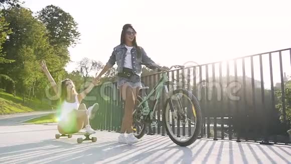 白种人年轻女性坐在长板板上而她最好的女性朋友在日落时骑着自行车走在她前面视频的预览图