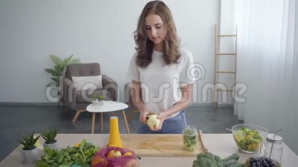 漂亮的年轻苗条的女人用锋利的刀在厨房的桌子旁剥苹果健康观念视频的预览图