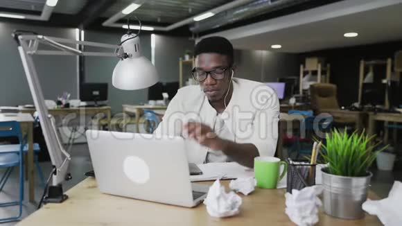 带着耳机的非洲年轻人正在笔记本电脑上工作在办公室里喝咖啡或茶视频的预览图