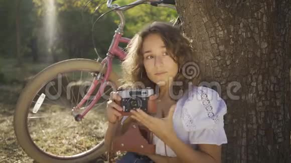 可爱的年轻摄影师卷发坐在树下用老相机在花园或公园里拍照视频的预览图