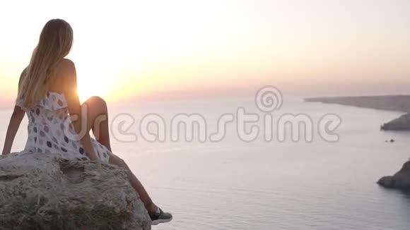 美丽的小女孩穿着漂亮的太阳裙坐在海边的岩石上欣赏着夕阳张开双手视频的预览图