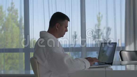 侧面观看自信的成年男子穿着白色外套坐在酒店房间的桌子上用笔记本电脑工作视频的预览图