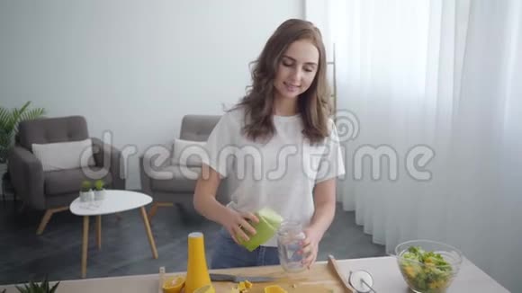 年轻苗条的女人在家里厨房的小罐子里从摇床上倒着冰沙健康食品的概念视频的预览图