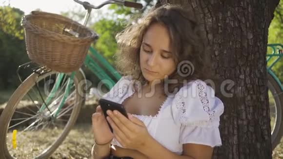 一位美丽的年轻女子卷发坐在树下她的手机在花园或公园里她的自行车视频的预览图