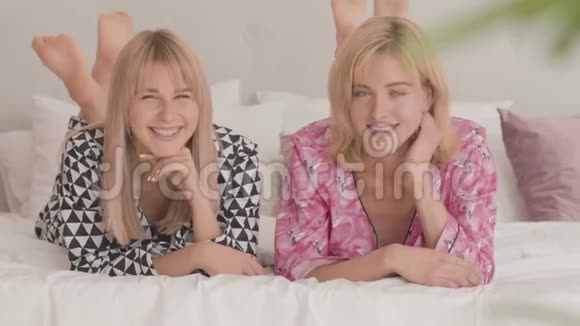 两个漂亮的金发白种人女孩穿着睡衣躺在白色柔软的床上看着相机快乐微笑的女人视频的预览图