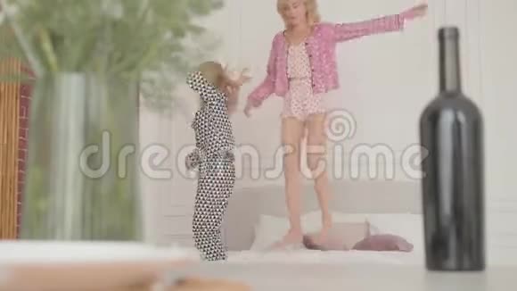 两个年轻的金发女孩在柔软的床上跳着笑快乐的朋友在家开睡衣派对重点是视频的预览图