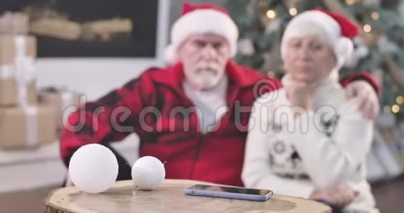 焦点从智能手机转变为坐在圣诞树旁边的悲伤的老夫妇的脸待召退休人员视频的预览图