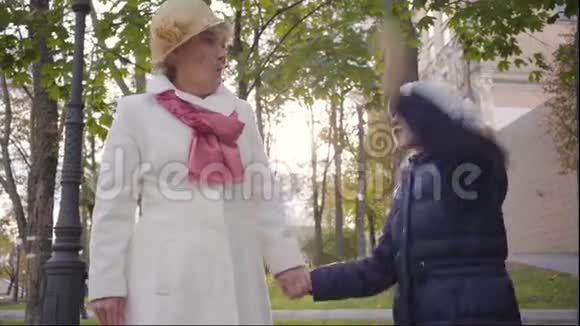 白种人成熟的女人戴着米色的科洛切帽子穿着白色外套和她开朗的孙女站在院子里视频的预览图