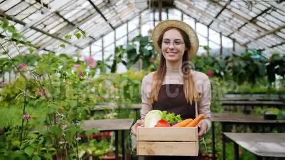 一个漂亮的金发女人带着一盒新鲜蔬菜在温室里散步视频的预览图