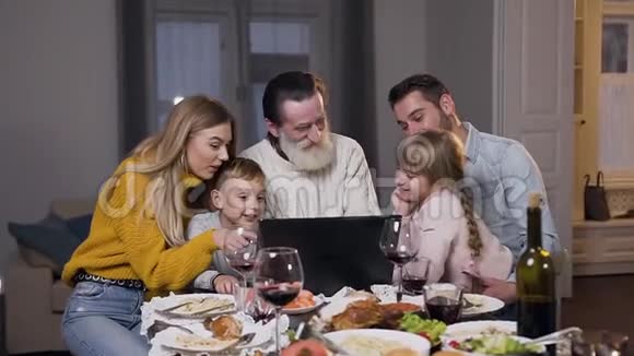 漂亮的不同的一代人坐在餐桌旁在笔记本电脑上看有趣的照片视频的预览图